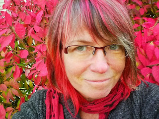 A White woman with glasses and multicolored bright pink strands among brown hair, chin length, in front of a background of pink flowers.