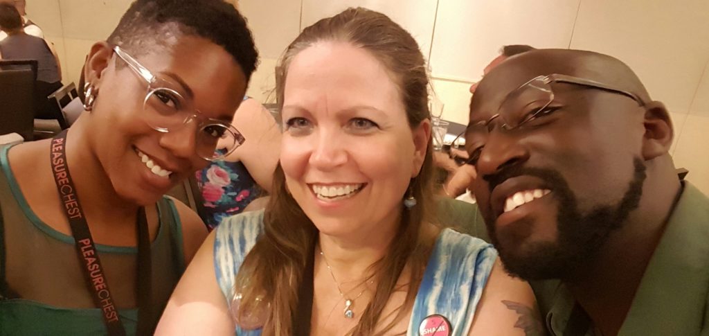 A beautiful Black woman with clear glasses and a handsome Black man with glasses and facial hair flank a white woman in a blue and white dress.