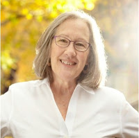 Smiling white woman with silver shoulder length hair and glasses, wearing a white shirt, with a background of golden trees