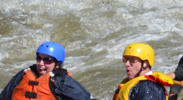 Two people wearing helmets, one smiling, with a swirl of white and green water behind them